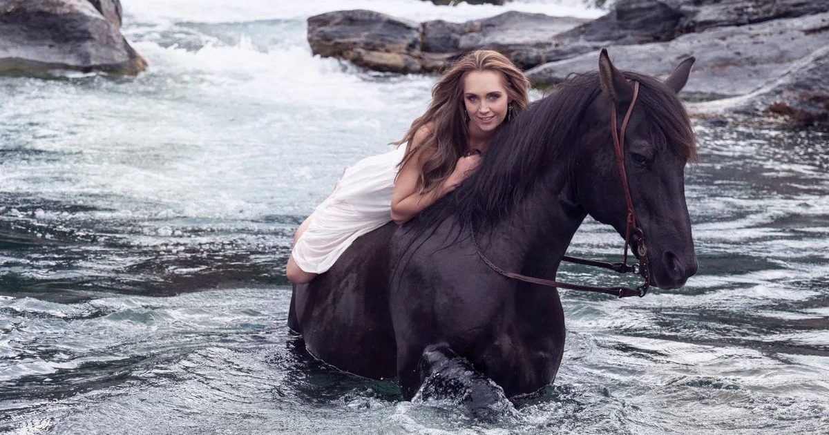 Amber Marshall riding her horse Hawk, who plays Spartan on Heartland, through a mountain river below a waterfall